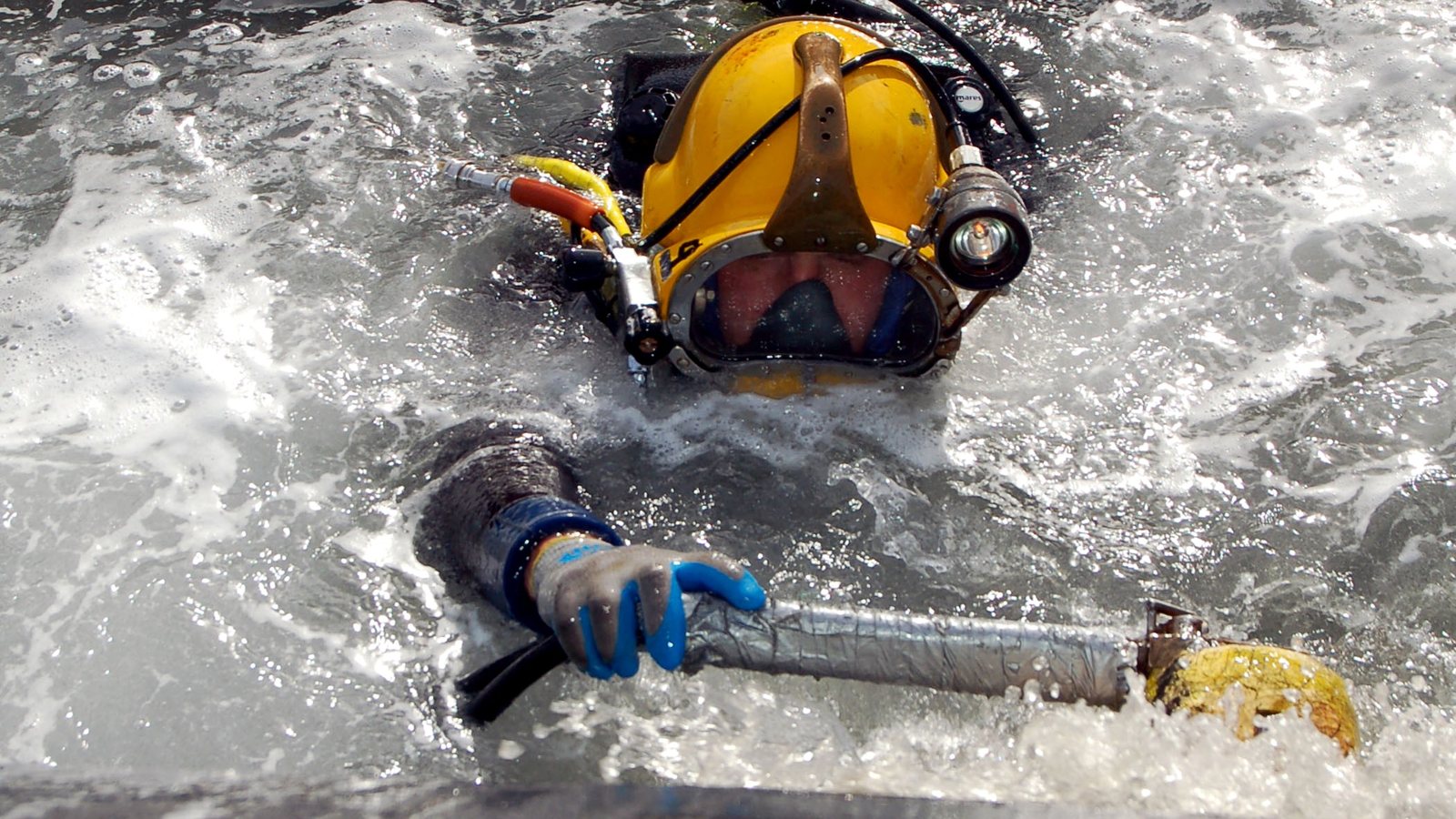 Diver in a surface-supplied helmet working a scrubber against a ship&rsquo;s hull at the waterline